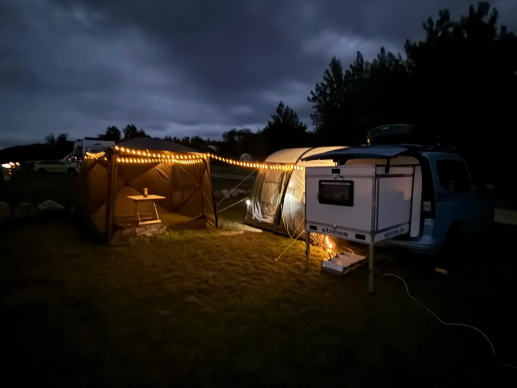 Blauer Van mit Ausziehmodul von ebicos.de, angeschlossenes Vorzelt und Pavillon mit Lichterkette bei Nacht – gemütliches Vanlife-Camping.