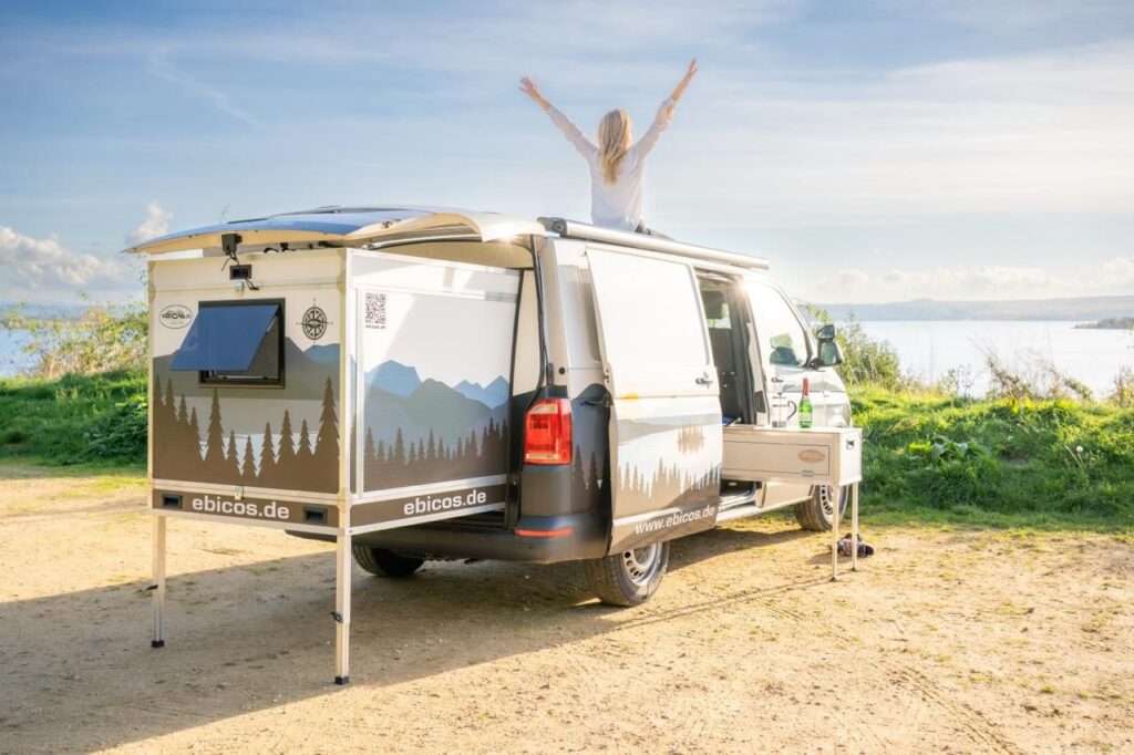 Van mit Happy Dreamer Ausziehmodul von ebicos.de – eine Frau genießt den Moment auf dem Dach mit Blick auf die Natur und den See. Perfekt für Vanlife und Campingabenteuer.
