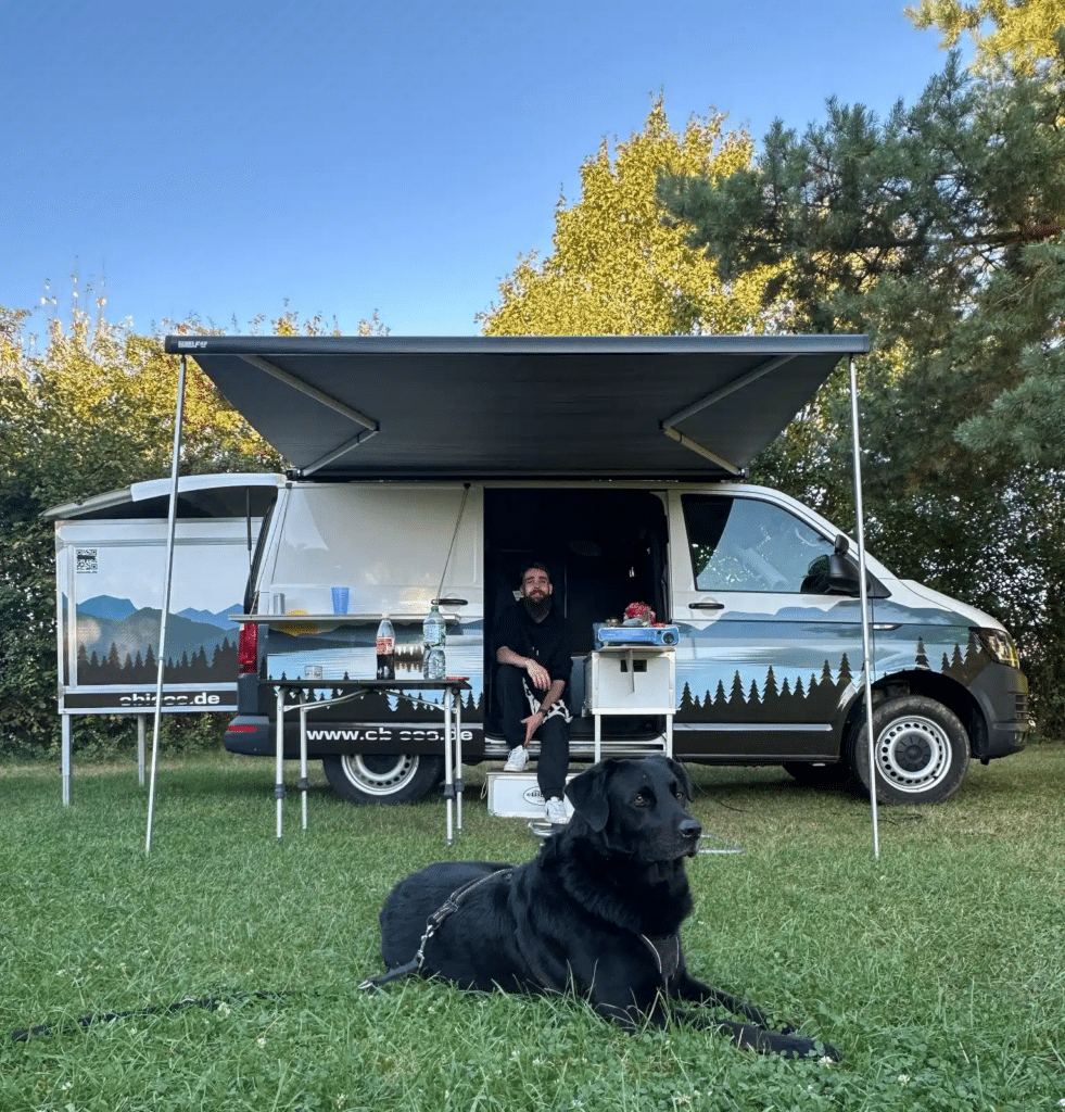 Van mit ausgefahrenem Happy Dreamer steht in der Natur. Herrchen und Hund sitzen gemütlich draußen und genießen das Wetter.