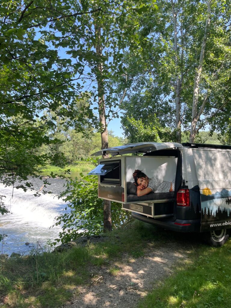 Ausgefahrenes Slide-Out-Schlafmodul Happy Dreamer Electric im Heck eines VW-Vans am Ufer der Neiße, Frau liegt auf der Matratze