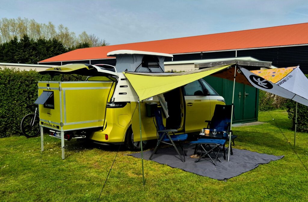 Gelber VW ID.Buzz mit ausgezogenem Schlafmodul und Sonnensegel auf einem Campingplatz