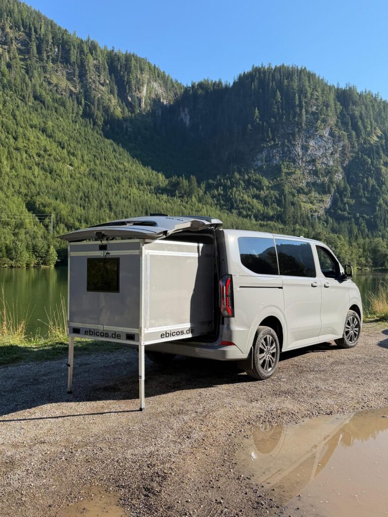 Weißer Ford Transit Custom mit ausgefahrenem Happy Dreamer Schlafmodul auf einem Schotterplatz am See in Österreich
